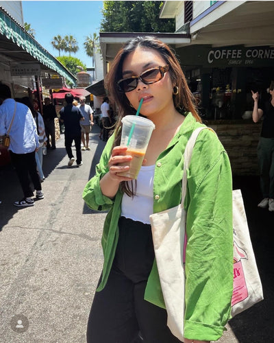 sunglasses, rectangle silhouette, tort frame, smokey brown lens, wide face sunglasses, wide face petite, slightly wider than standard, midi sunglasses, mini sunglasses, slightly smaller than standard - shown worn by a female with long brown hair, standing in an outdoor market, looking toward the camera, with a coffee cup in one hand and has it raised to her lips, and a canvas tote over her other sholder, she is wearing green shirt over white tanktop and black pants