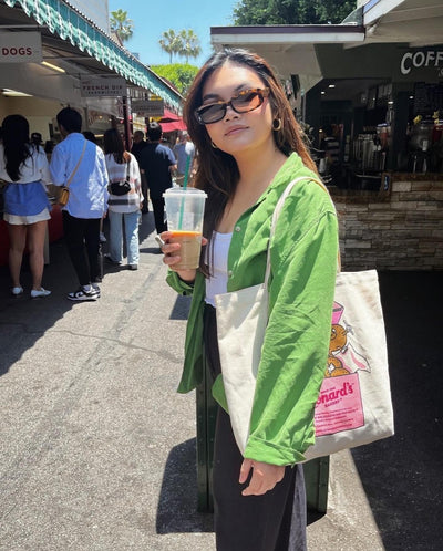 sunglasses, rectangle silhouette, tort frame, smokey brown lens, wide face sunglasses, wide face petite, slightly wider than standard, midi sunglasses, mini sunglasses, slightly smaller than standard - shown worn by a female with long brown hair, standing in an outdoor market, looking toward the camera, with a coffee cup in one hand, and a canvas tote over her other sholder, she is wearing green shirt over white tanktop and black pants