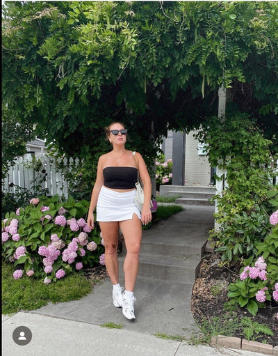 sunglasses, trapezoid silhouette, trap silhouette, black frame, midi size, wide face petite, slightly wider than standard - shown as worn on a woman wearing a black strapless top, white skirt, and white sneakers, she stands below an archway of vines surrounded by gardens with flowering bushes, and a concrete path behind her