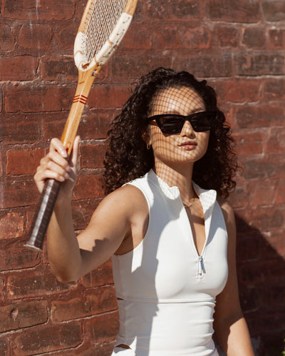 sunglasses, trapezoid silhouette, trap silhouette, black frame, midi size, wide face petite, slightly wider than standard - shown on a women with curly brown hair, wearing a white tennis outfit, she is holding a tennis racquet up toward the sun casting a shadow on her face, she stands by a red brick wall