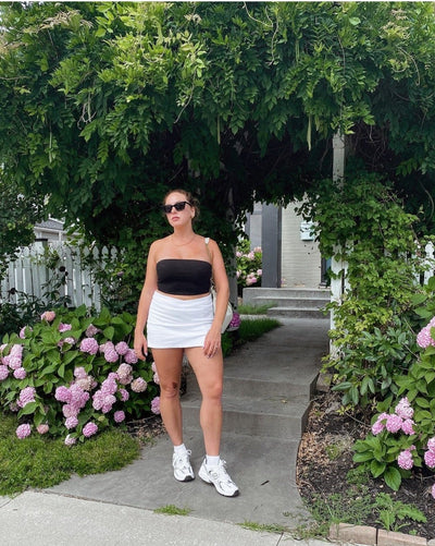 sunglasses, trapezoid silhouette, trap silhouette, black frame, midi size, wide face petite, slightly wider than standard - shown as worn on a woman wearing a black strapless top, white skirt, and white sneakers, she stands below an archway of vines surrounded by gardens with flowering bushes, and a concrete path behind her