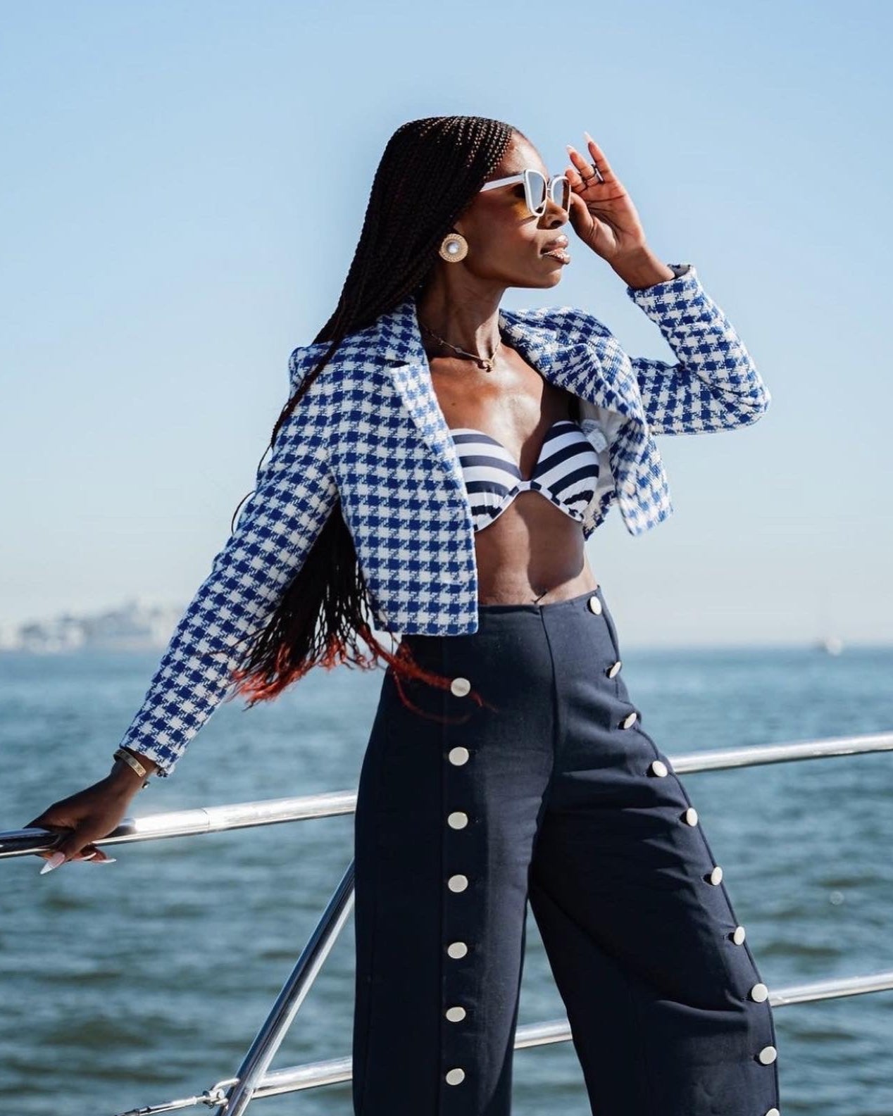 Model standing in front of a railing near water, wearing white Sol Kyst cat eye sunglasses and checkered attire. 