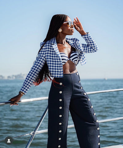 sunglasses, kyst eye, cat eye silhouette, white frame, smokey lens, wide face sunglasses - shown as worn by a women with rich brown skin, long waist length hair in braids, she is on a boat, standing with the water behind her, she is wearing a blue and white striped bikini top, blue and white cropped blazer, and navy blue pants with white button detail down the front of each leg