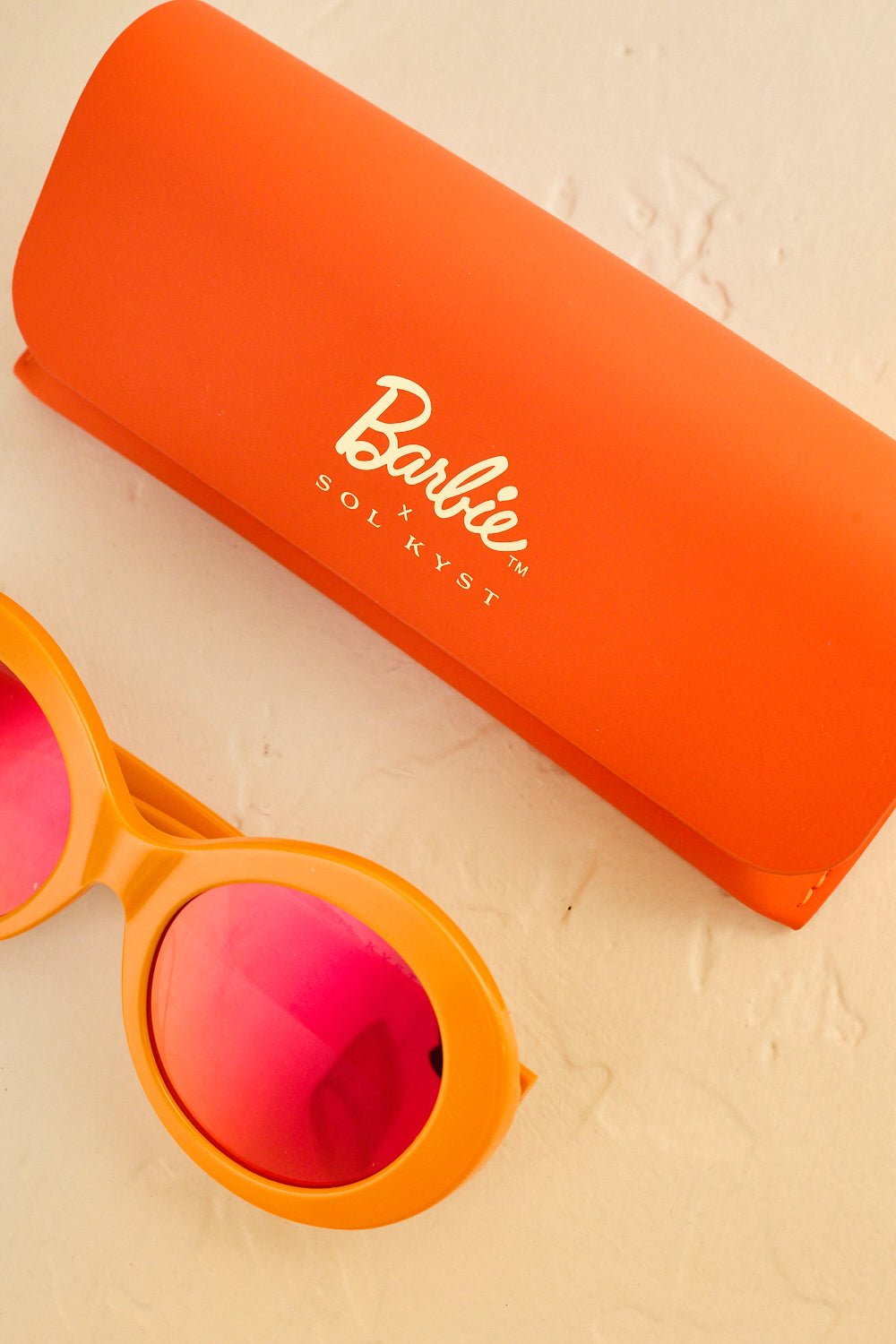 Orange Barbie Sol Kyst sunglasses with pink-tinted lenses, displayed alongside an orange case on a light textured surface.