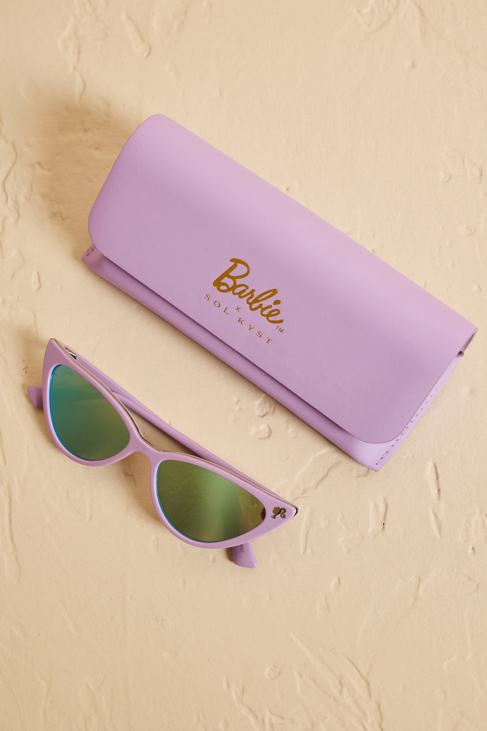 Barbie x Sol Kyst lavender cat eye sunglasses with matching branded case on a beige surface - top view.