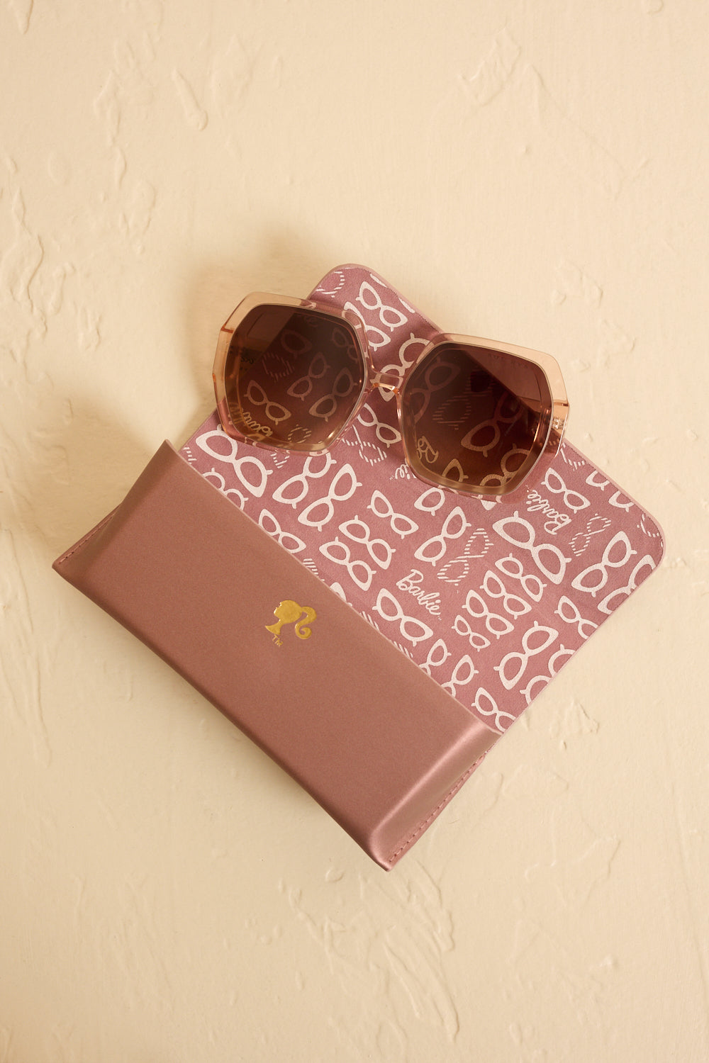 Hexagonal sunglasses with brown translucent frames and brown lenses, placed on an open Barbie x Sol Kyst case with a patterned interior.