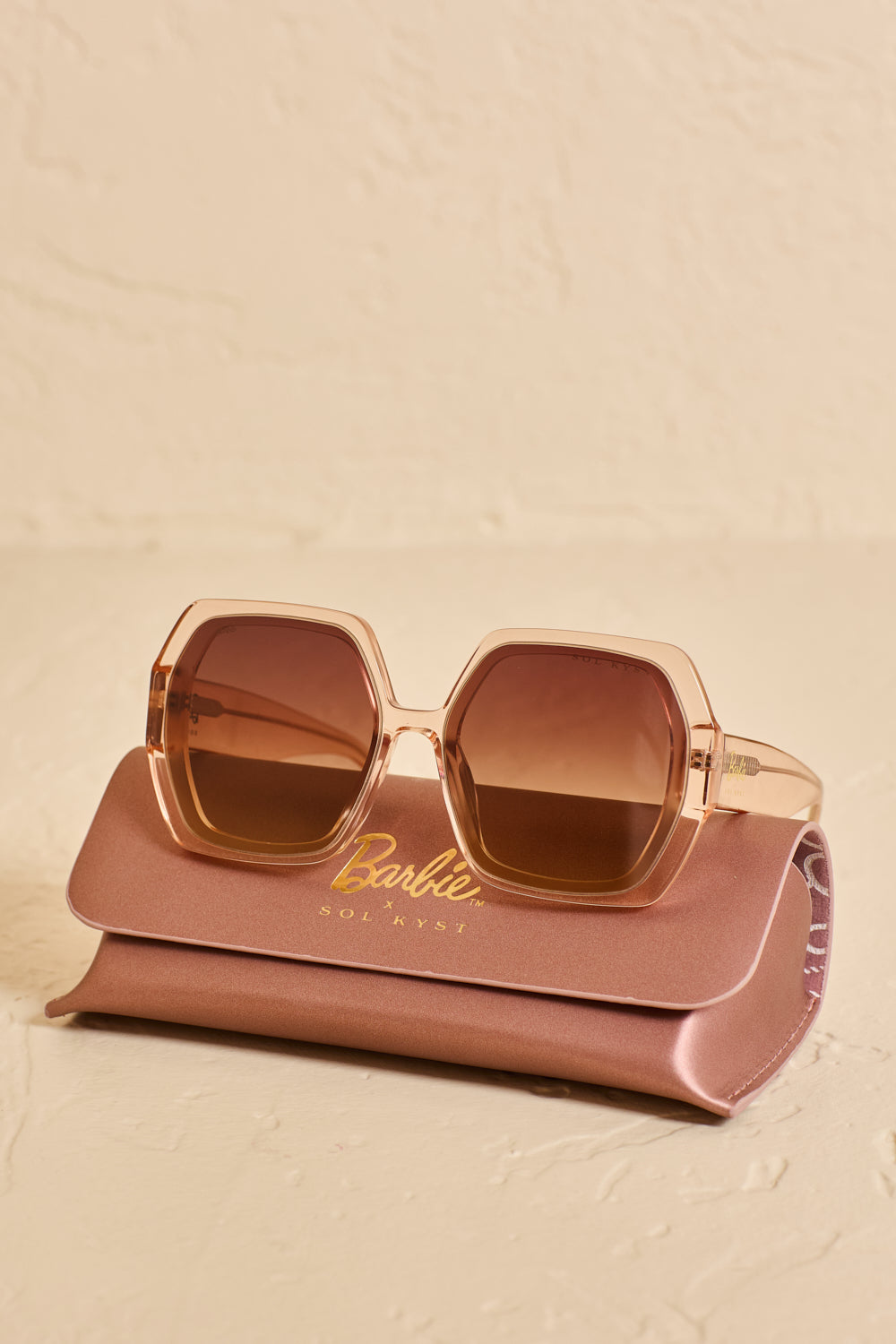 Barbie x Sol Kyst hexagonal sunglasses with brown translucent frames and brown lenses, displayed on matching case on a beige surface.