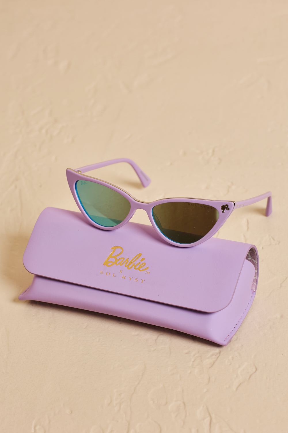 Barbie x Sol Kyst lavender cat eye sunglasses with reflective lenses, on top of a matching branded case on a beige surface.