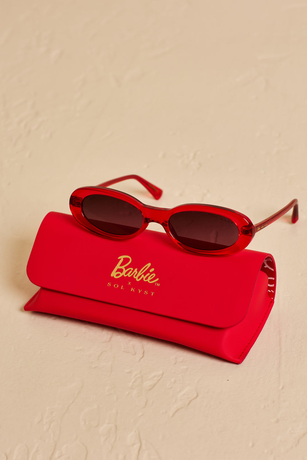 Barbie x Sol Kyst red oval sunglasses displayed with a matching red case on a beige textured surface.