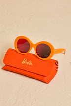 Barbie Sol Kyst round orange sunglasses with red-tinted lenses, displayed on top of an orange case on a textured beige surface.