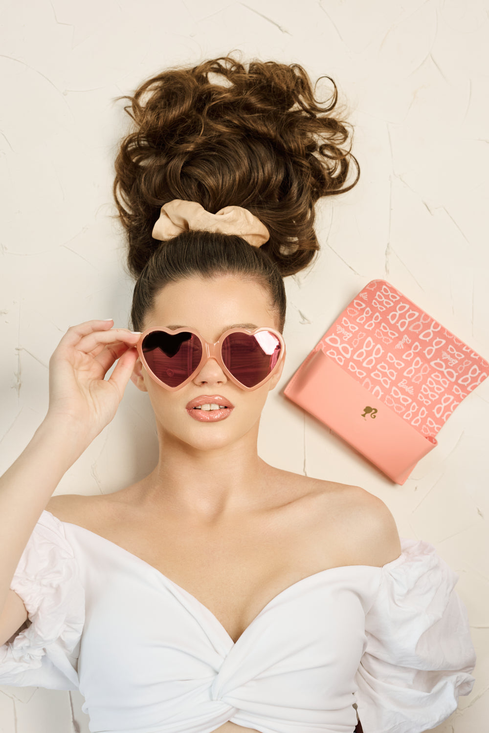 Woman wearing peach-pink Barbie x Sol Kyst heart-shaped sunglasses, with matching case in the background.
