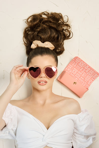 Woman wearing peach-pink Barbie x Sol Kyst heart-shaped sunglasses, with matching case in the background.