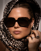 Woman wearing sunglasses and a leopard print headscarf with a neutral background