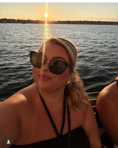 sunglasses, kyst eye, cats eye silhouette, black frame, wide face sunglasses - shown as worn by a female sitting on a boat, with the water and setting sun behind her, she is wearing a black halter neck stop and large gold earrings