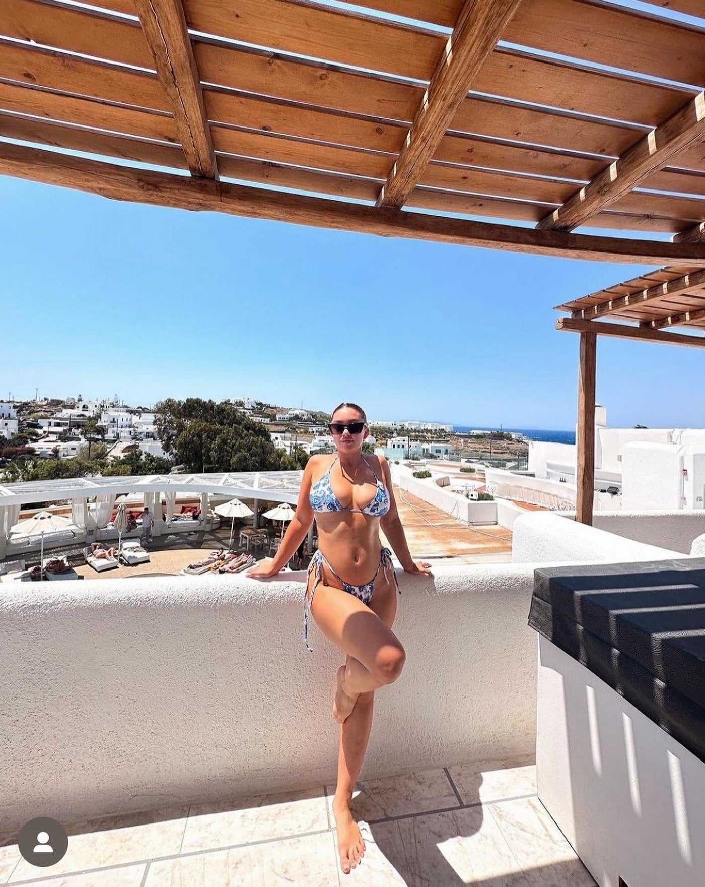 sunglasses, trapezoid silhouette, trap silhouette, black frame, midi size, wide face petite, slightly wider than standard - shown being worn by a women with hair pulled back, wearing a blue and white paisley print bikini, standing below a wood pergola, on a balcony with a white stucco wall behind her, beyond her is blue sky, white buildings, trees, and lounge chairs around a pool below the balcony 