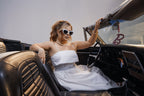 Model sitting in retro car wearing Barbie cat eye sunglasses and a white dress.