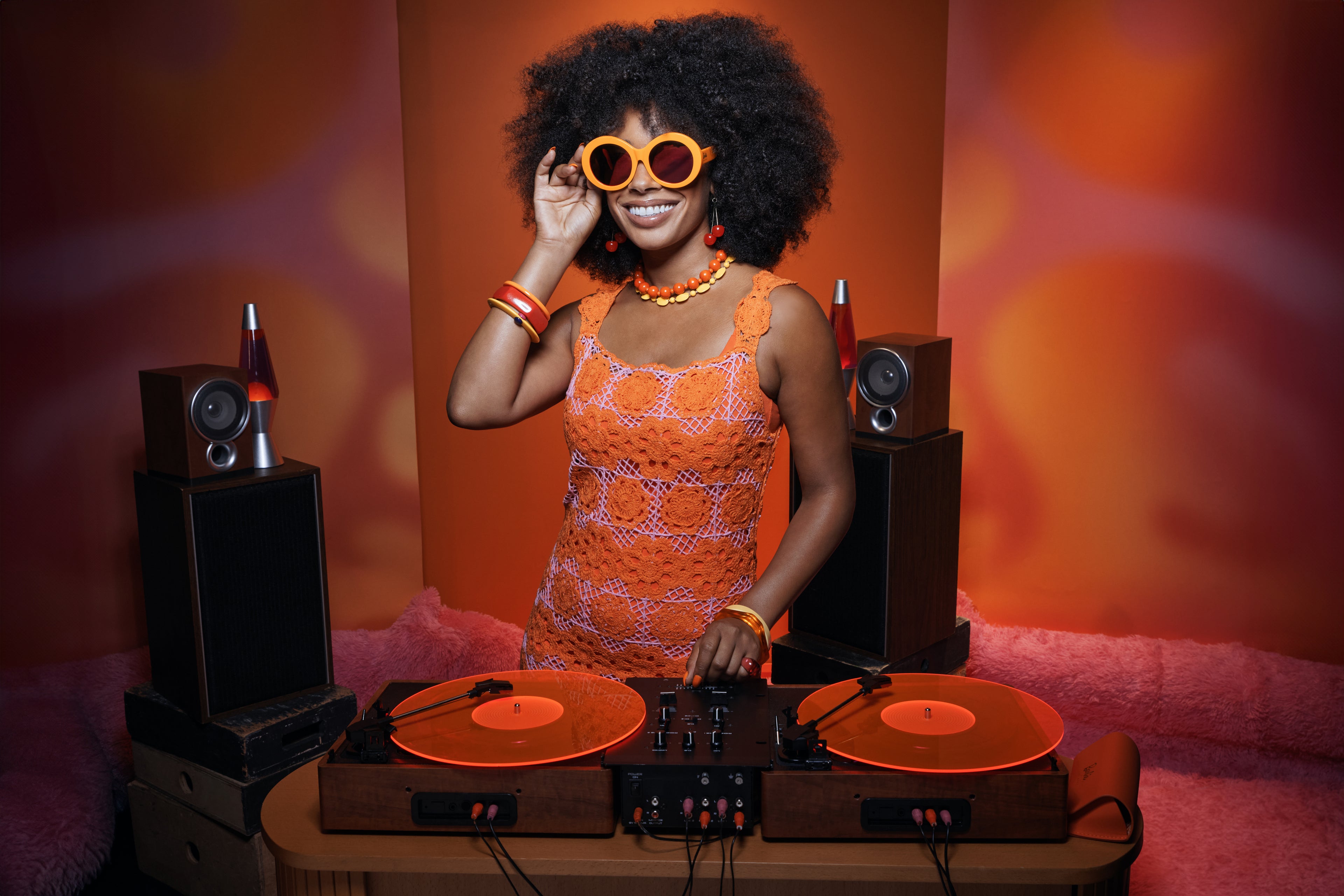 Woman wearing Barbie Sol Kyst orange sunglasses with pink-tinted lenses, standing by a DJ turntable in a retro orange setup.