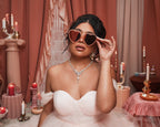 Woman wearing Barbie x Sol Kyst heart-shaped sunglasses, styled with an elegant necklace and pink decor backdrop.