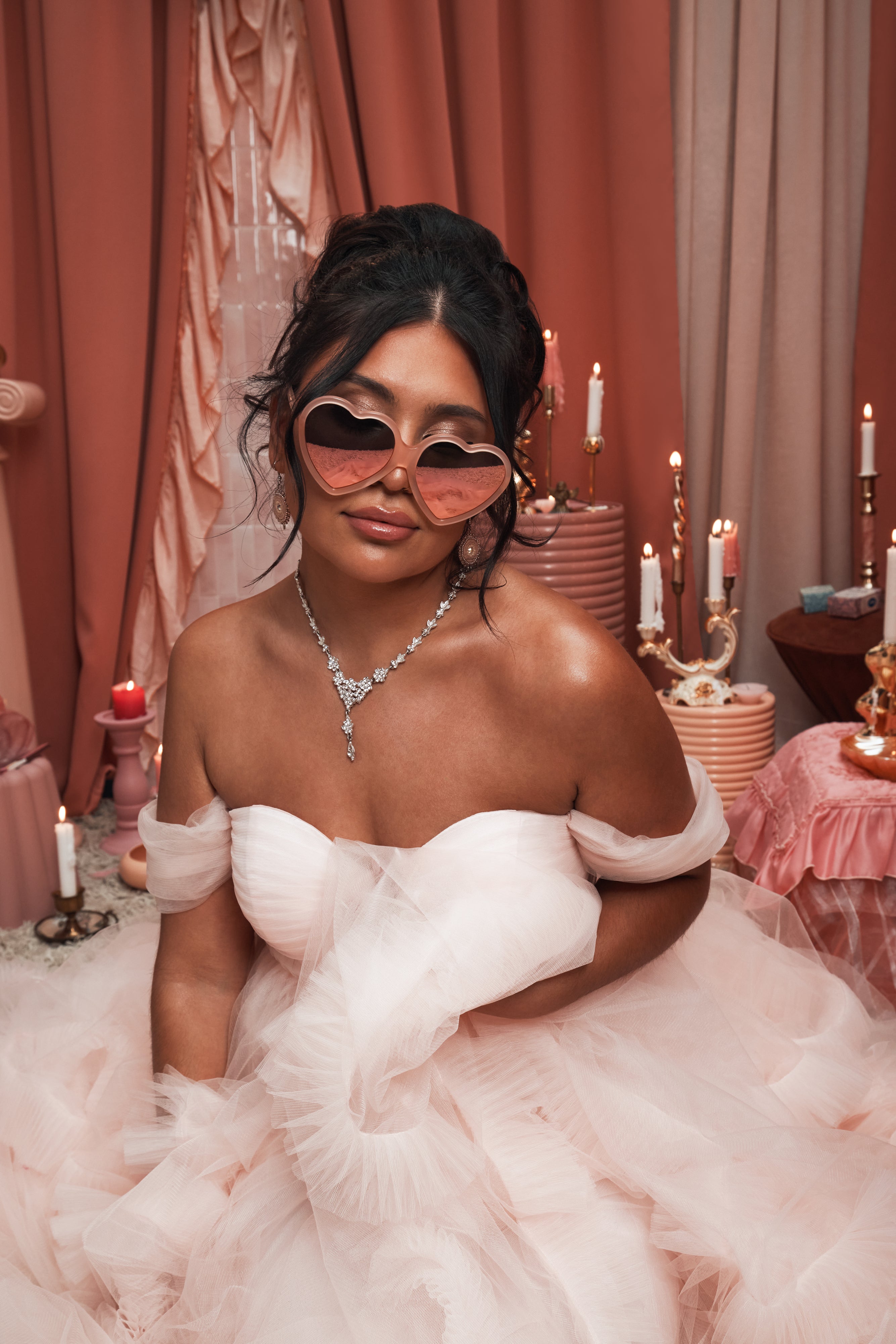 Woman in a soft pink tulle gown wearing Barbie x Sol Kyst heart-shaped sunglasses, surrounded by peach-toned decor and candles.