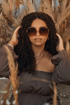 Model with brown curly hair and matching dress wearing Barbie x Sol Kyst brown hexagonal sunglasses.