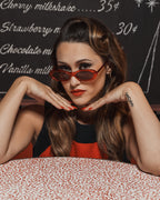 Woman wearing Barbie red oval sunglasses, posing at a retro diner table with vintage decor.