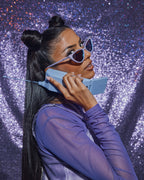 Model dressed with lavender Barbie cat-eye sunglasses, a vintage blue phone, and a glittering purple background.