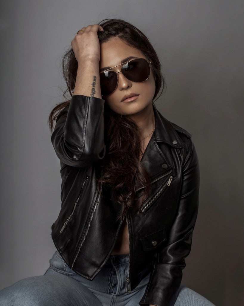 sunglasses, aviator silhouette, wide face sunglasses, gold metal frame, mirrored lens, black ear piece - shown as worn on a female with long dark brown hair, she is wearing a black leather jacket and light blue jeans, she is sitting, with one arm raised up and her hand in her hair
