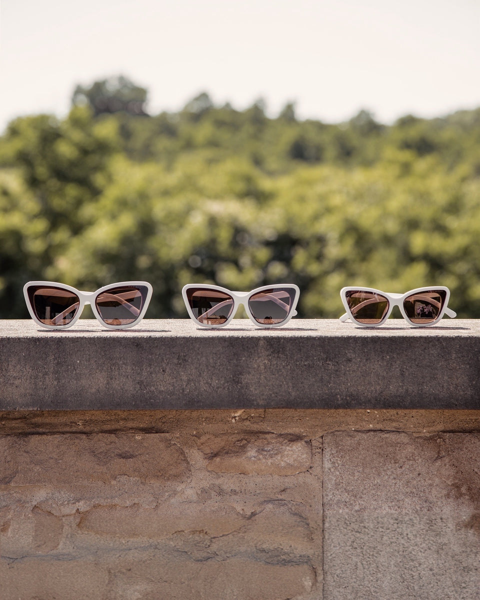 Three Sol Kyst Bridal Collection white cat-eye sunglasses displayed on a stone ledge, featuring white frames and brown lenses.
