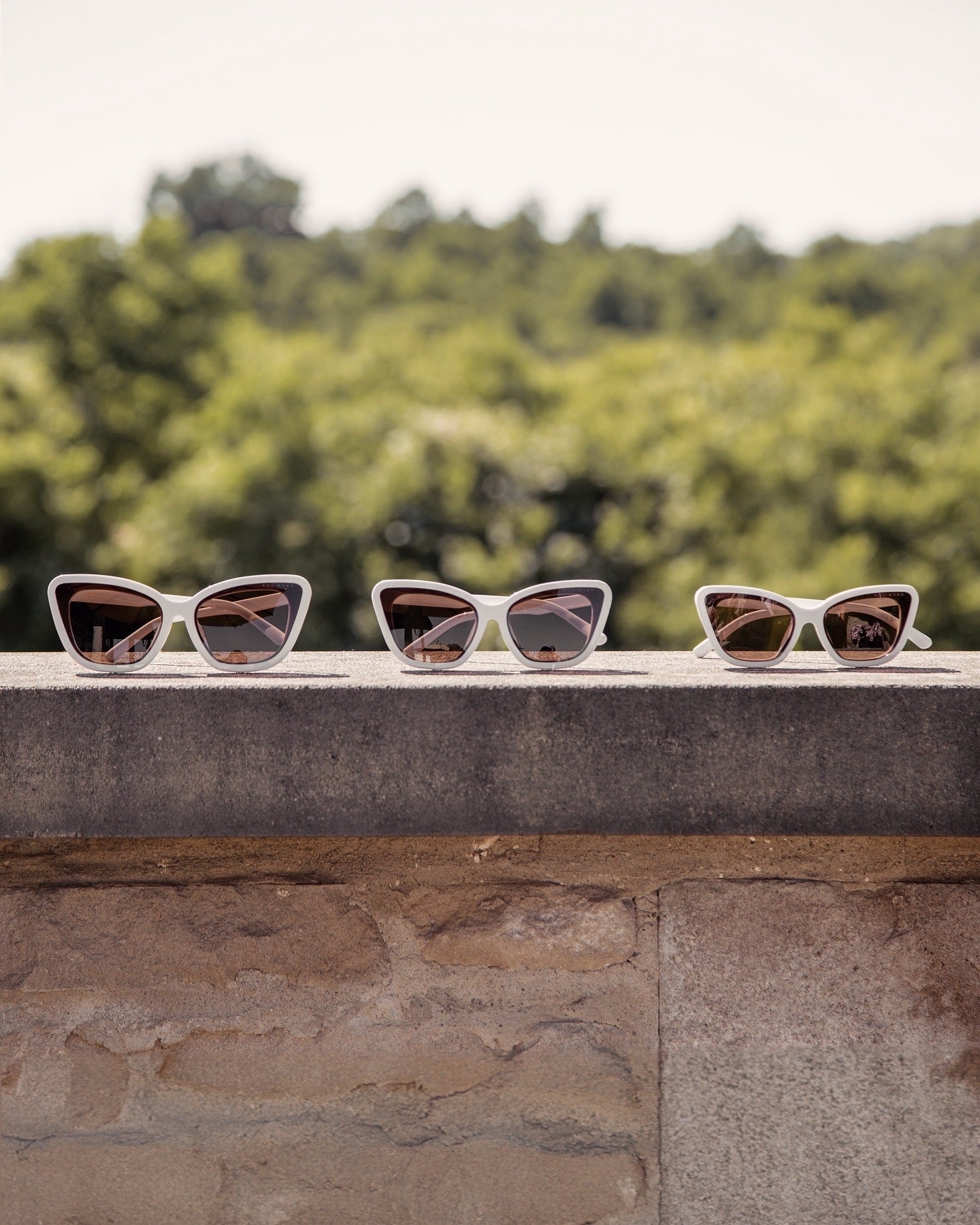 Three Sol Kyst Bridal Collection white cat-eye sunglasses displayed on a stone ledge, featuring white frames and brown lenses.