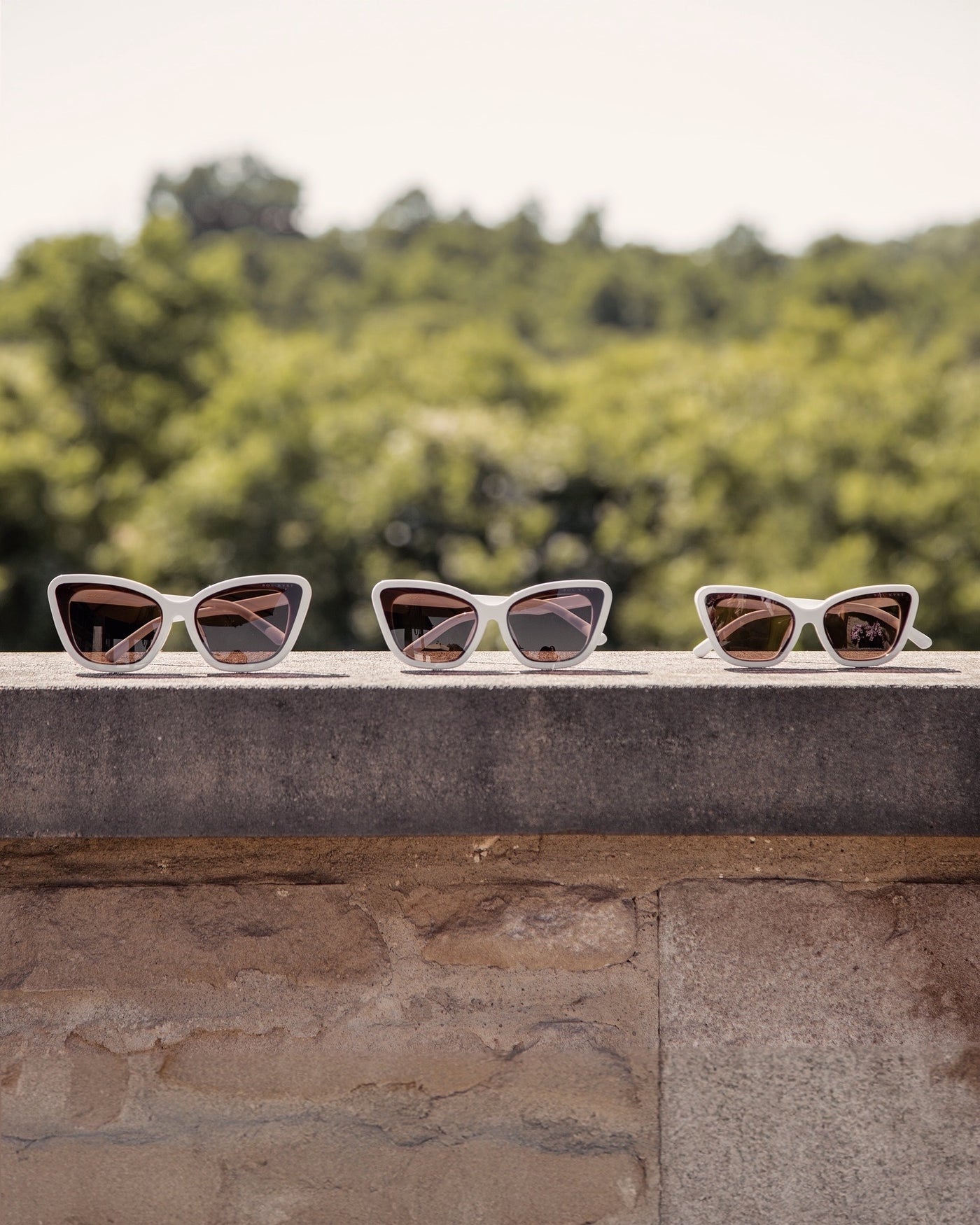 Three Sol Kyst Bridal Collection white cat-eye sunglasses displayed on a stone ledge, featuring white frames and brown lenses.
