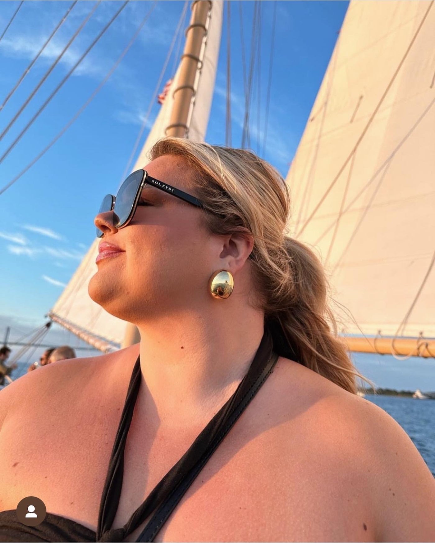 sunglasses, kyst eye, cats eye silhouette, black frame, wide face sunglasses - shown as worn by a female sitting on a boat, with the sails behind her, she is looking out over the water,, she is wearing a black halter neck stop and large gold earrings