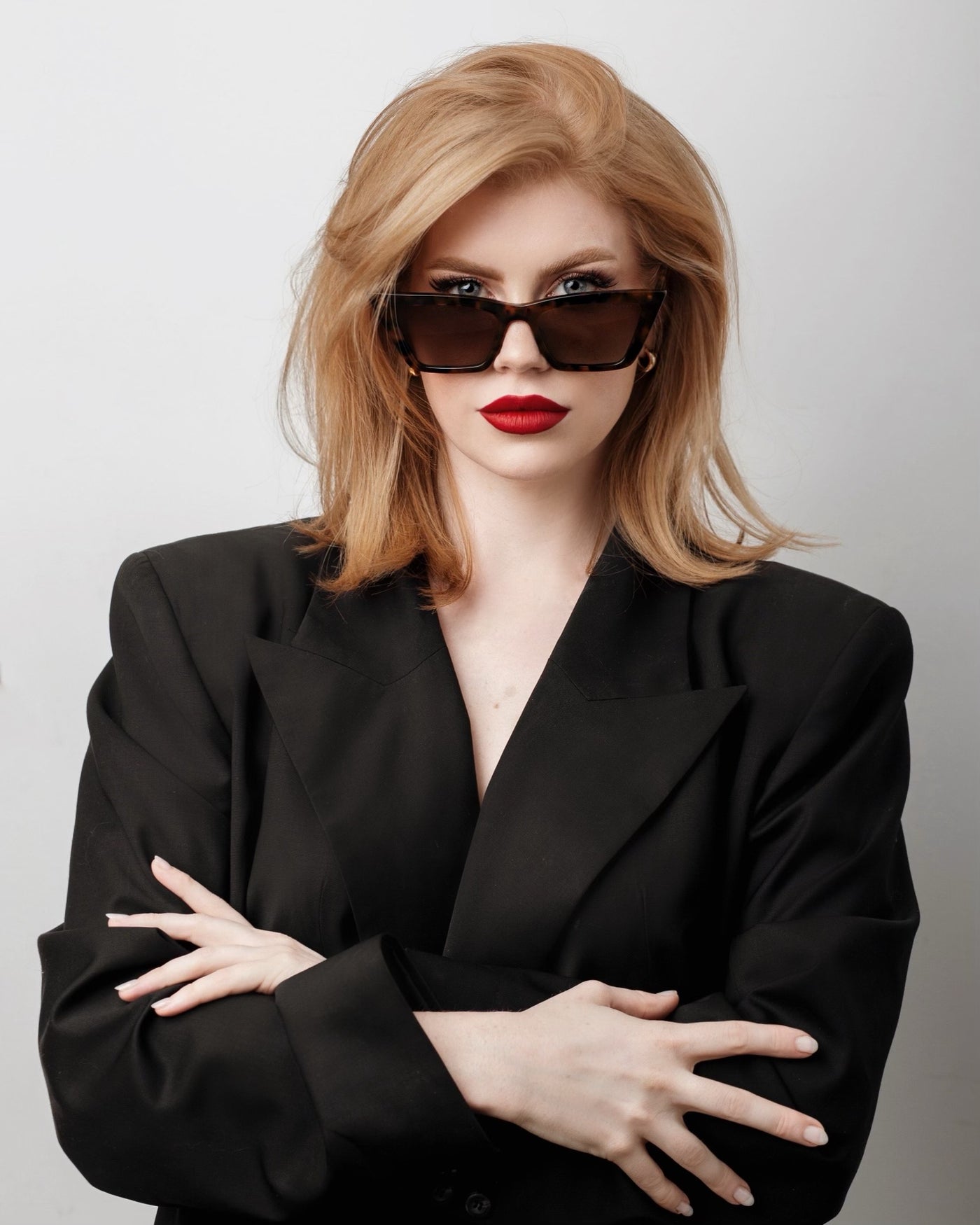 sunglasses, Trapezoid silhouette, trap silhouette, Tort frame, smokey brown lens, wide face petite, midi sunglasses, slightly wider than standard - shown as worn by female with orange-y red hair, dark red lipstick, black blazer, gold jewelry, she is looking toward the camera, arms crossed with hands on arms, glasses sitting down the bridge of her nose so you can see her eyes