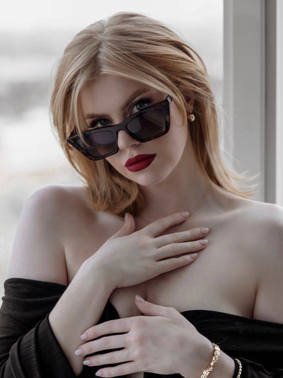 sunglasses, Trapezoid silhouette, trap silhouette, Tort frame, smokey brown lens, wide face petite, midi sunglasses, slightly wider than standard - shown as worn by female with orange-y red hair, dark red lipstick, black off the shoulder dress, gold jewelry, she is looking toward the camera with glasses slightly down the bridge of her nose so you can see her eyes, her hands are on her chest