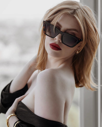 sunglasses, Trapezoid silhouette, trap silhouette, Tort frame, smokey brown lens, wide face petite, midi sunglasses, slightly wider than standard - shown as worn by female with orange-y red hair, dark red lipstick, black off the shoulder dress, gold jewelry, she is looking toward the camera