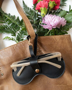 Sol Kyst black Kyst Klip holding transparent sunglasses, attached to a brown bag with red and pink flowers in the background.