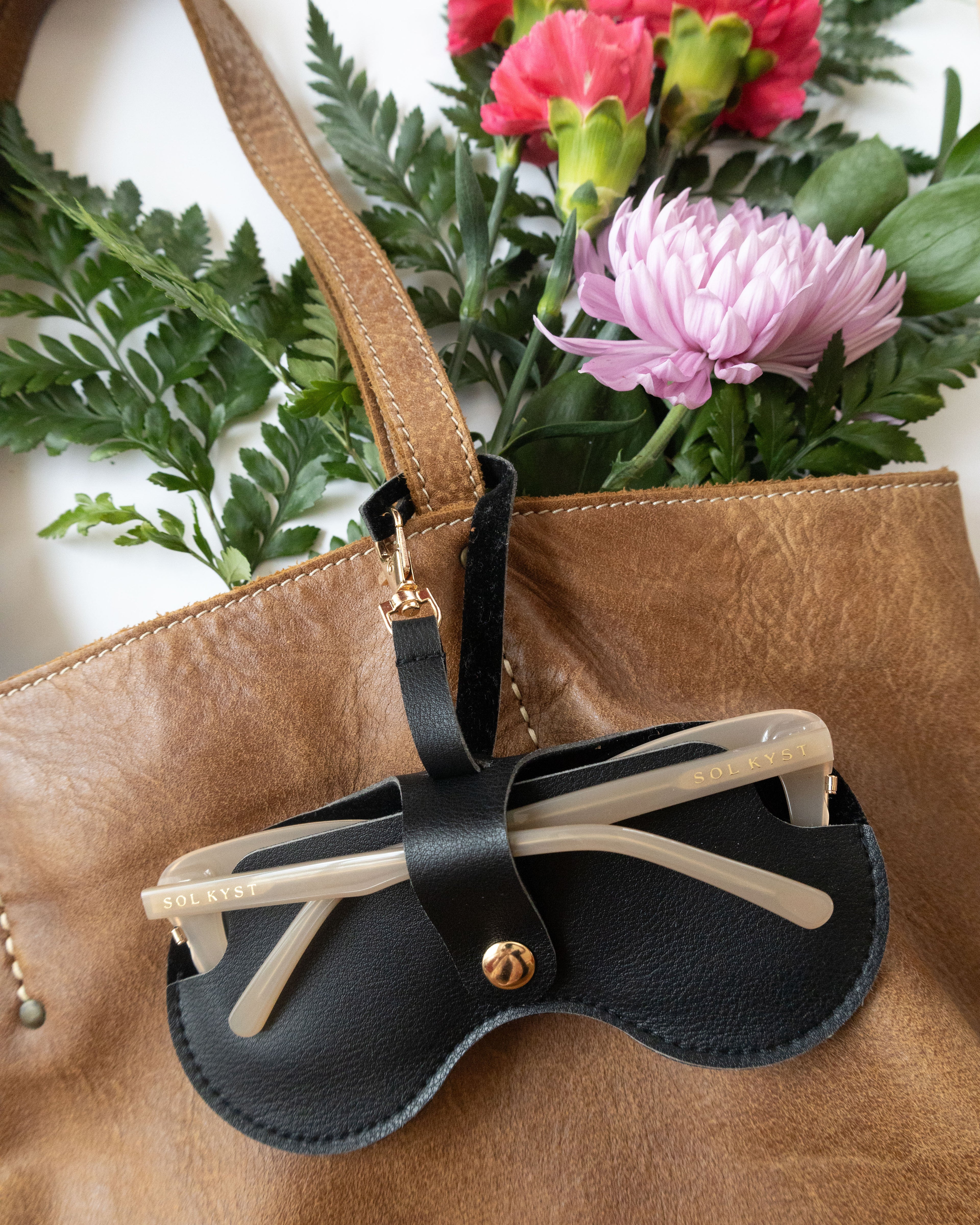 Sol Kyst black Kyst Klip holding transparent sunglasses, attached to a brown bag with red and pink flowers in the background.