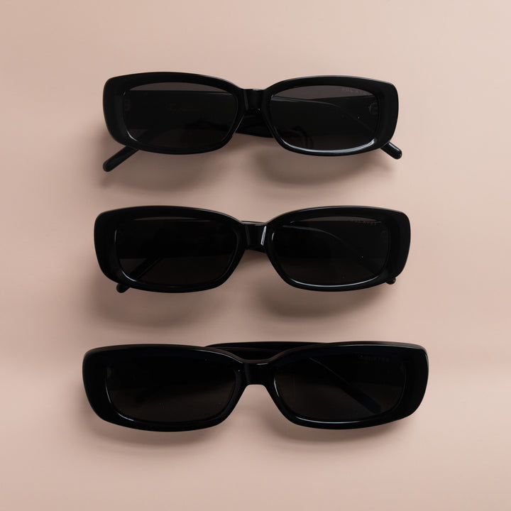 Three pairs of rectangular black sunglasses with dark tinted lenses arranged vertically on a beige surface.