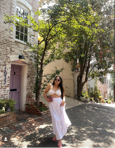 sunglasses, kyst eye, cat eye silhouette, white frame, smokey lens, wide face sunglasses - shown as worn by a women with long dark brown hair, standing on the street line with brick houses and trees, she is wearing all white, holding a small purse