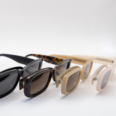 Four rectangular sunglasses in black, tortoise, beige, and cream frames with tinted lenses, displayed in a row at an angle on a white surface.