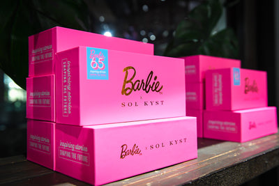 Barbie x Sol Kyst pink boxes with gold logos, stacked on a wooden display under dim lighting.