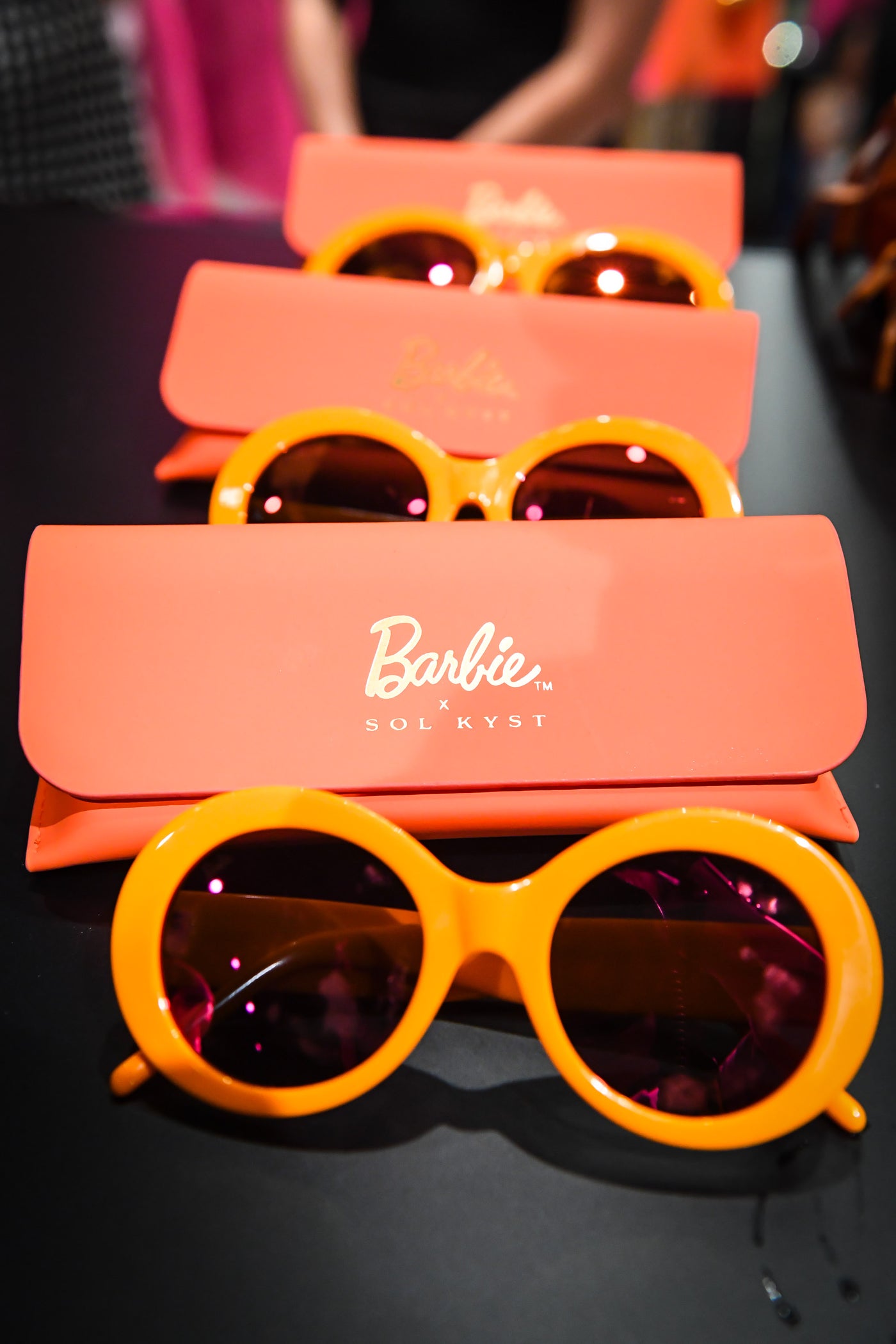 Three pairs of Barbie Sol Kyst round sunglasses with orange frames in a row, each with a matching case.