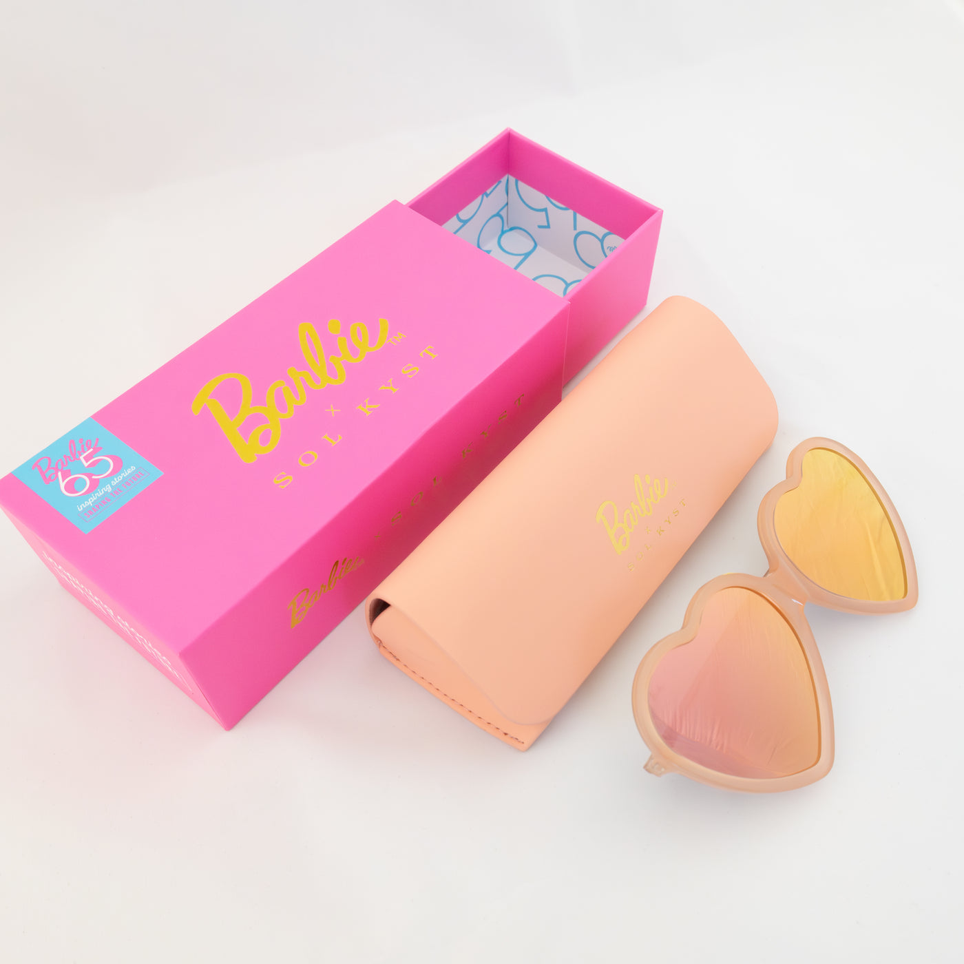 Barbie x Sol Kyst heart-shaped sunglasses with peach-pink frames, matching case, and vibrant pink packaging with gold branding.