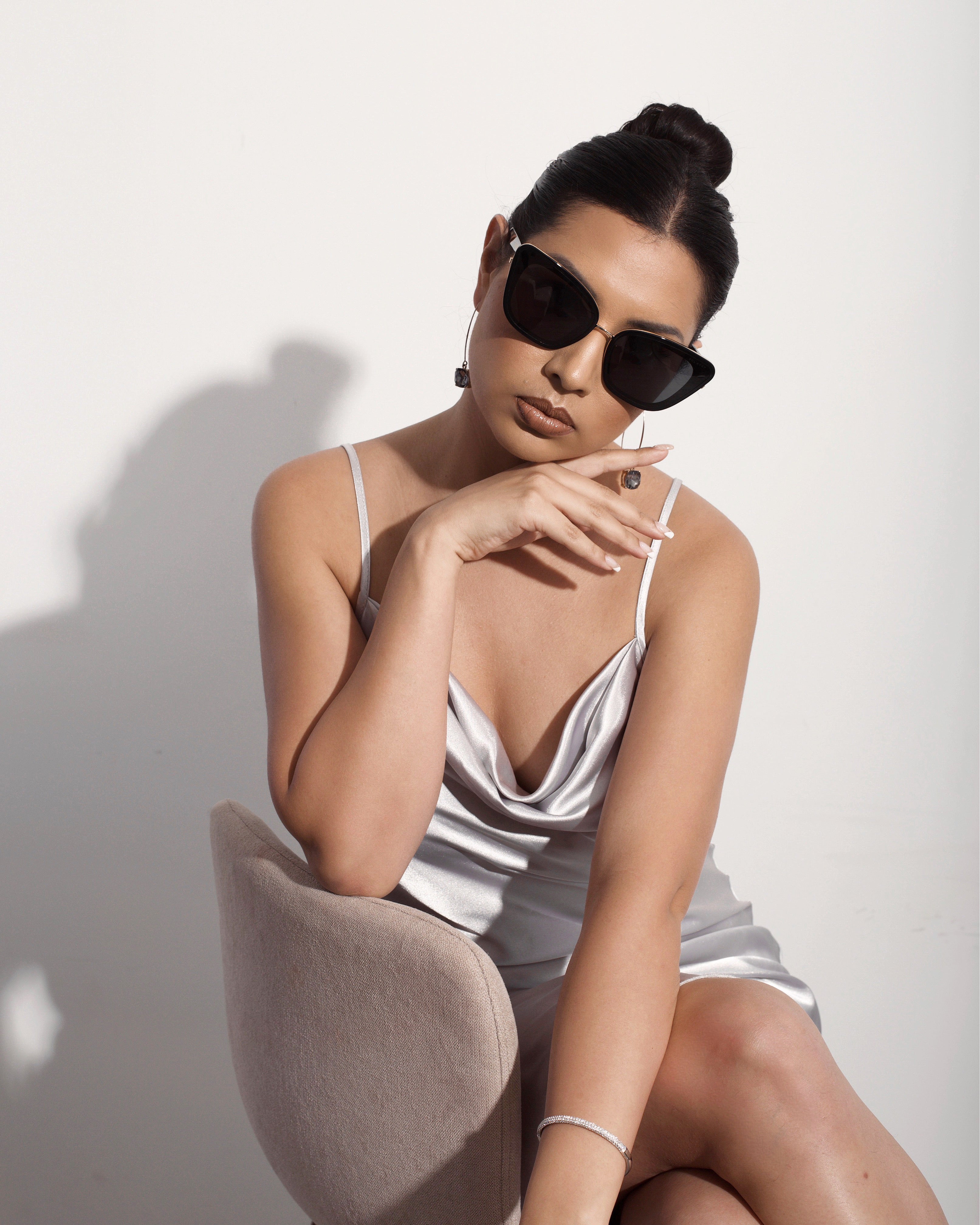 Model in satin gray dress sitting on a chair wearing Sol Kyst cat eye sunglasses with tinted lenses.
