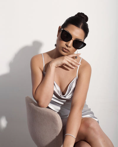 sunglasses, kyst eye, cats eye silhouette, black frame, wide face sunglasses - shown as worn by a women with dark hair pulled back in a bun, she is wearing a silver slip dress, and is posed sitting on a chair with one elbow resting on the back of the chair and that hand up below her chin