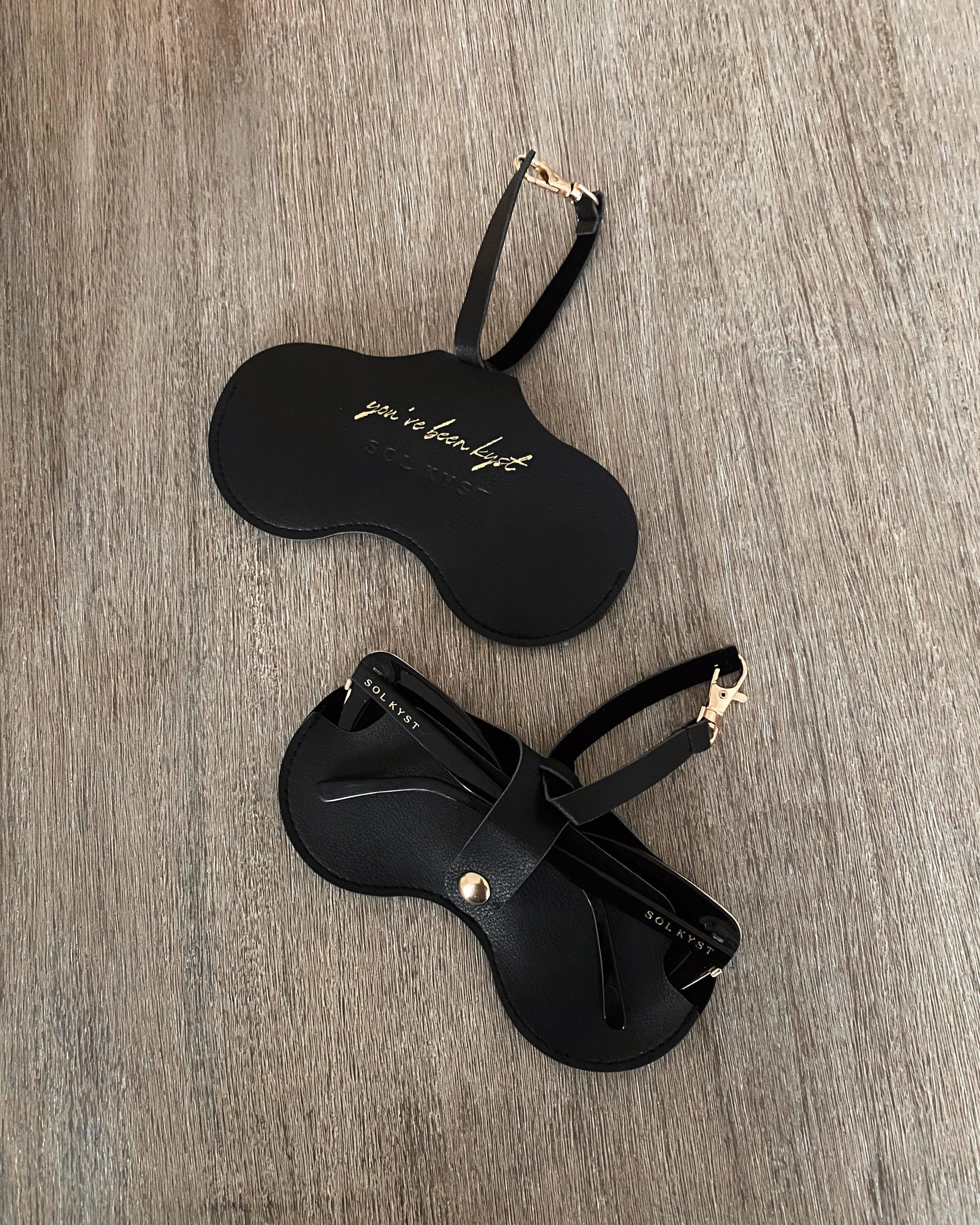 Black Sol Kyst Kyst Klip shown in two views: one with gold 'you’ve been kyst' text and another holding sunglasses, placed on a textured wooden surface.
