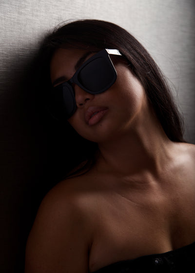 sunglasses, wayfarer silhouette, black frame, black lens, wide face sunglasses, wide face petite, midi size, slightly wider than standard - shown worn on a female, long dark brown hair, she is in the shadows, wearing a black strapless, 