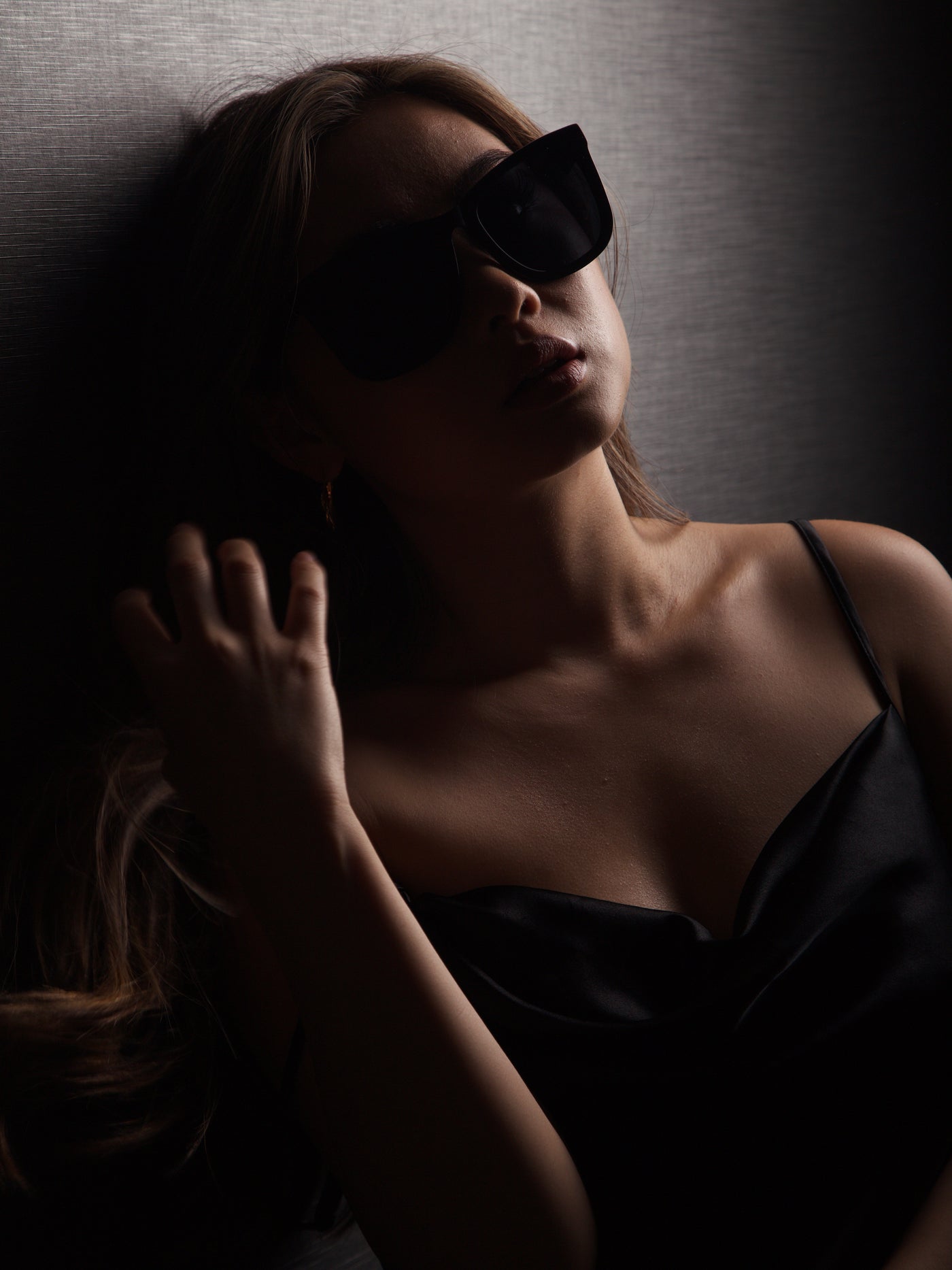 sunglasses, wayfarer silhouette, black frame, black lens, wide face sunglasses, wide face petite, midi size, slightly wider than standard - shown worn on a female, long light brown hair, she is in the shadows, wearing a black dress, with one hand raised to her hair