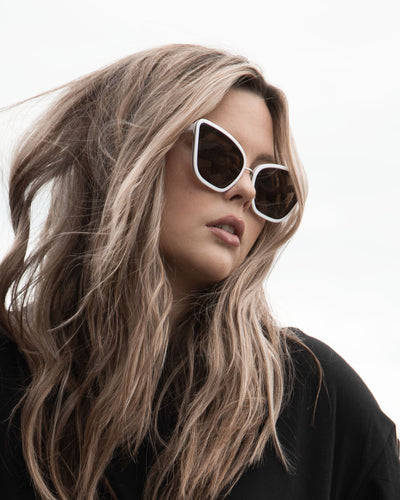 sunglasses, kyst eye, cat eye silhouette, white frame, smokey lens, wide face sunglasses, shown worn by a women with long blond wavy hair, wearing a black dress