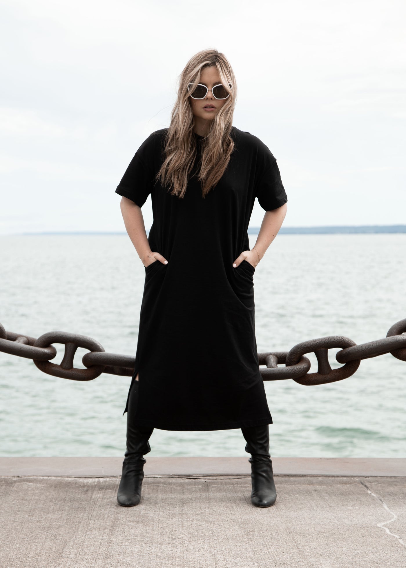 sunglasses, kyst eye, cat eye silhouette, white frame, smokey lens, wide face sunglasses - shown on woman standing facing camera, she has long blond hair, is wearing a long black short sleeve dress and black boots, her hands are in the pockets of her dress, she is standing in front of a large body of water on a path with a thick chain link hanging behind her 