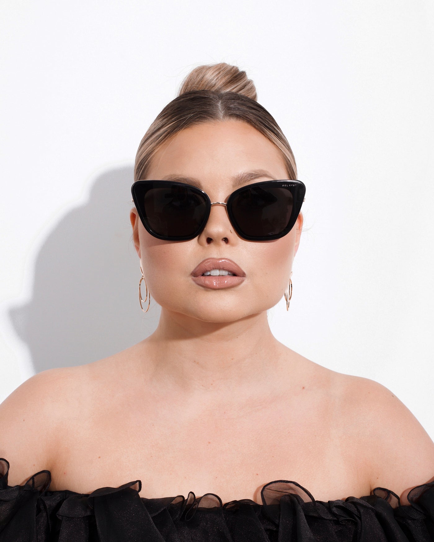 sunglasses, kyst eye, cats eye silhouette, black frame, wide face sunglasses, shown as worn by a woman wearing her blond hair tied back in a slick bun, gold earrings, and a black ruffled off the shoulder top